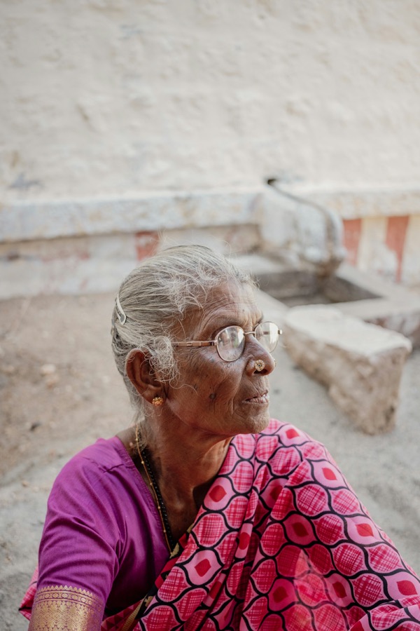 An elderly Tamil woman with glasses sitting outside her home