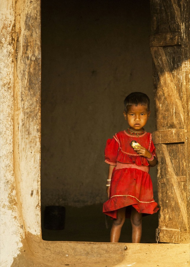 A young child in a red dress standing in the doorway of a mud home